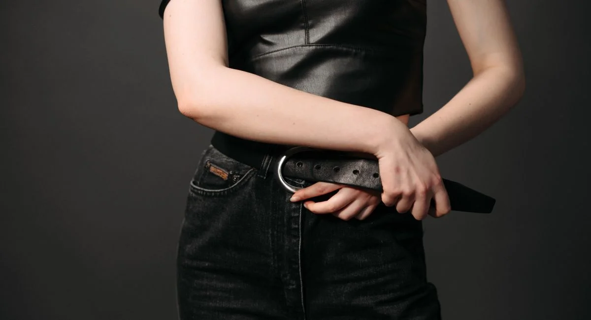 Stylish close-up of woman in black leather top and jeans holding a belt, showcasing modern fashion.