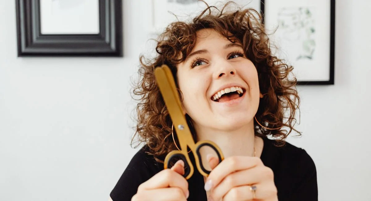 A cheerful woman with curly hair smiling while holding scissors, indoors.