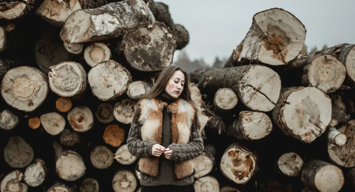 Stylish woman in a fur vest poses by stacked wood logs, evoking a rustic woodland feel.