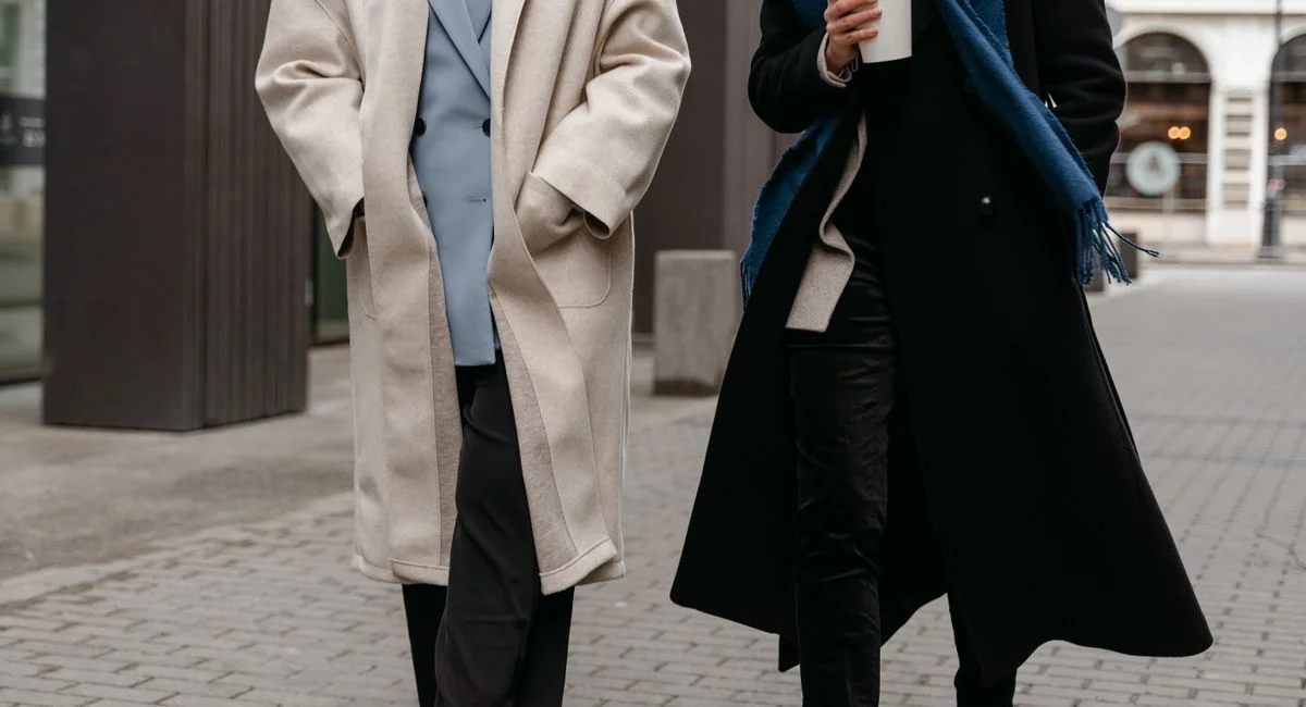 Two adults in stylish coats walk on a city street, enjoying a coffee break.