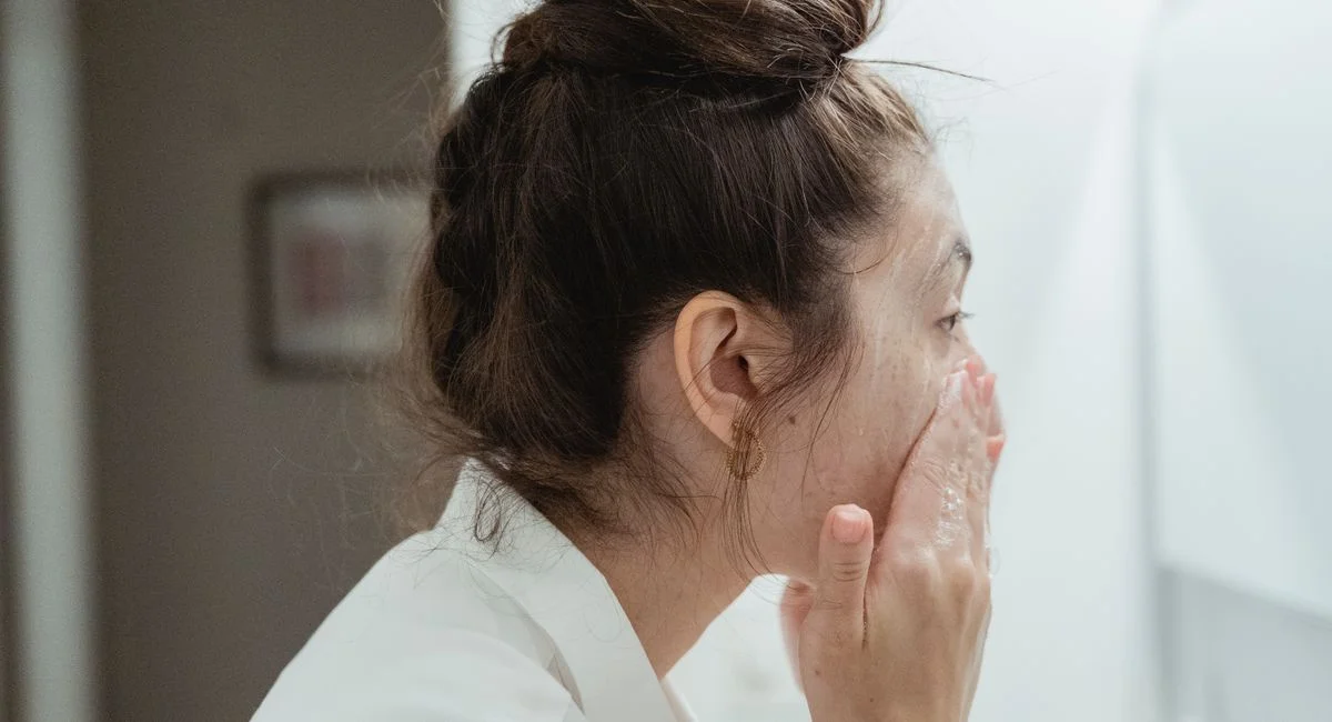 A young woman in a bathrobe follows her skincare routine, washing her face in a bathroom.