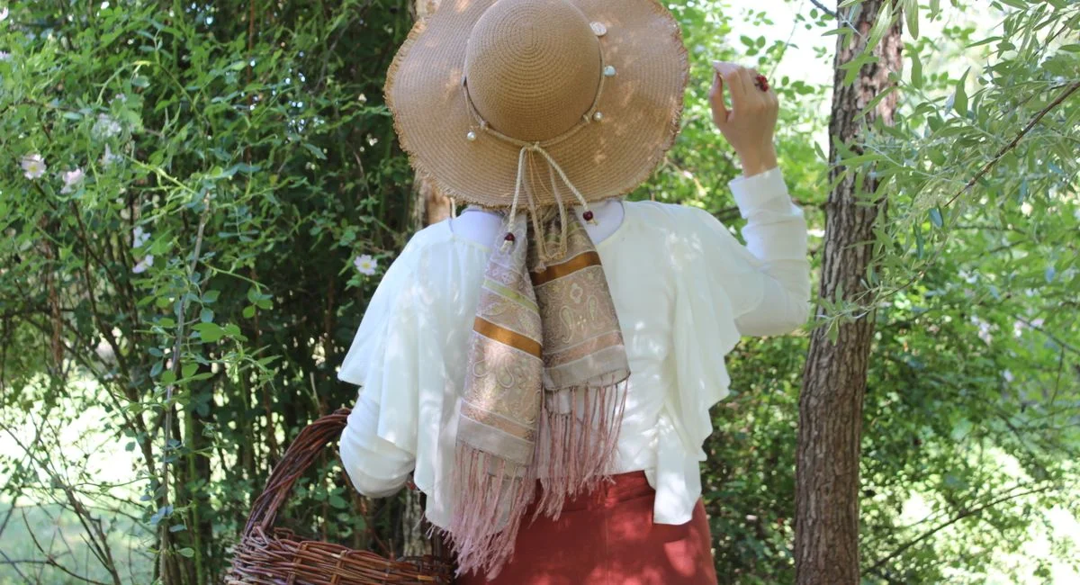 A woman wearing a straw hat and scarf holds a basket in a summer garden, surrounded by greenery.
