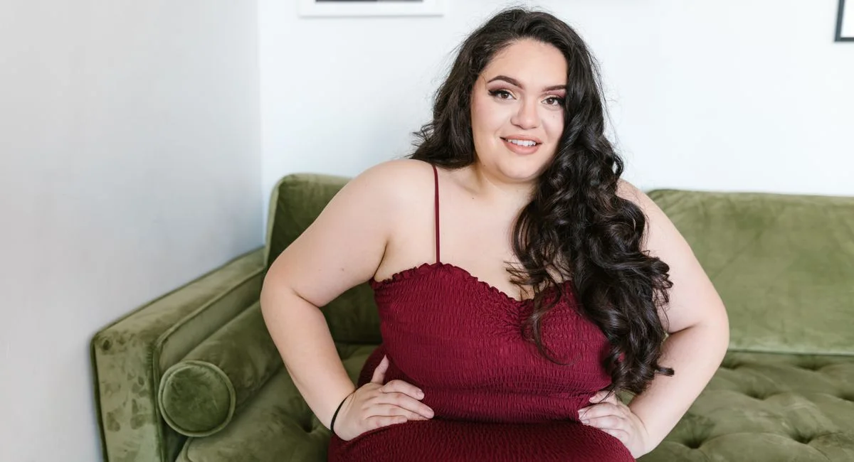 Confident plus-size woman in red dress sitting on a green sofa, embracing body positivity.