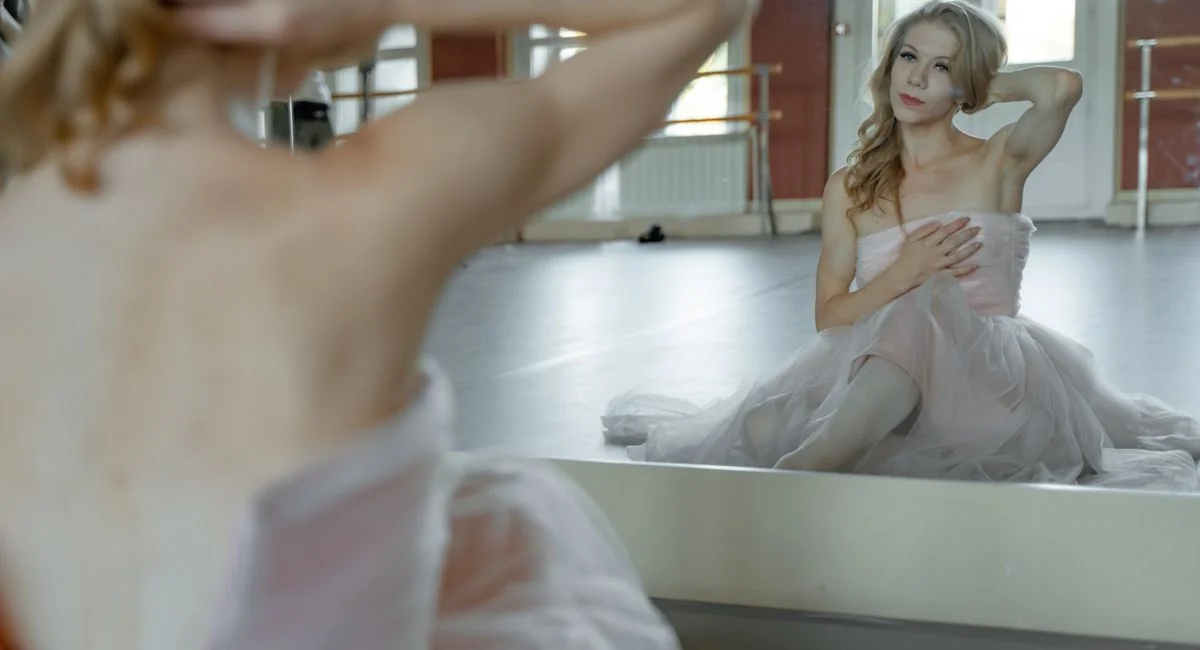 A ballet dancer in a pink dress poses in front of a mirror in a dance studio.