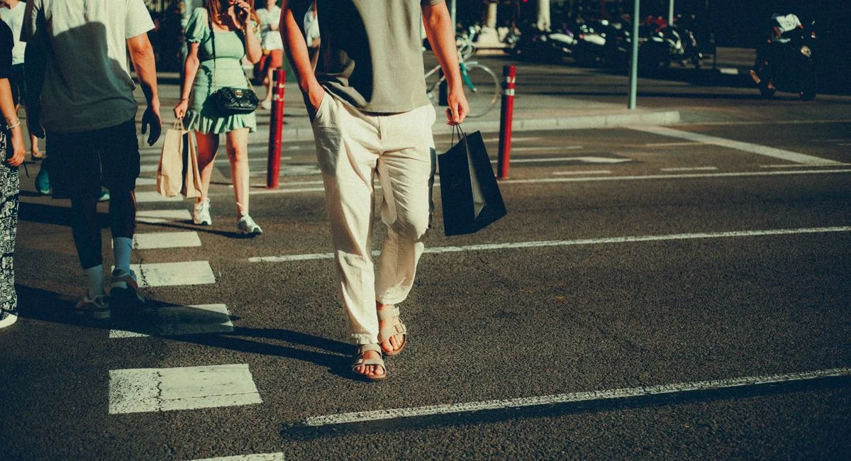 People walking across a sunlit city street in casual attire, holding shopping bags.